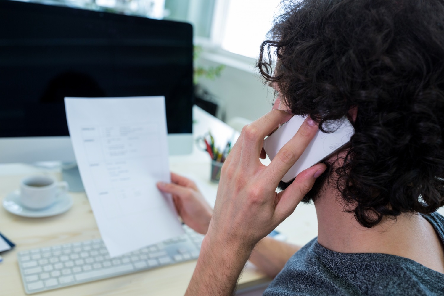 A student sitting in front of a computer, calling someone on his cellphone while holding a sheet of paper.